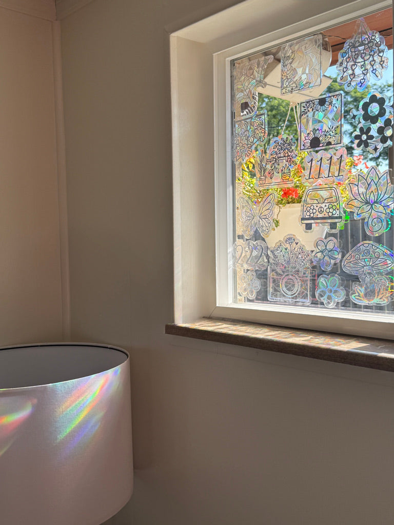 A window in a room decorated with many colorful, patterned, and glittery stickers and decals. In the bottom-left corner, a white cylindrical lampshade is visible, reflecting a rainbow of light onto the wall.