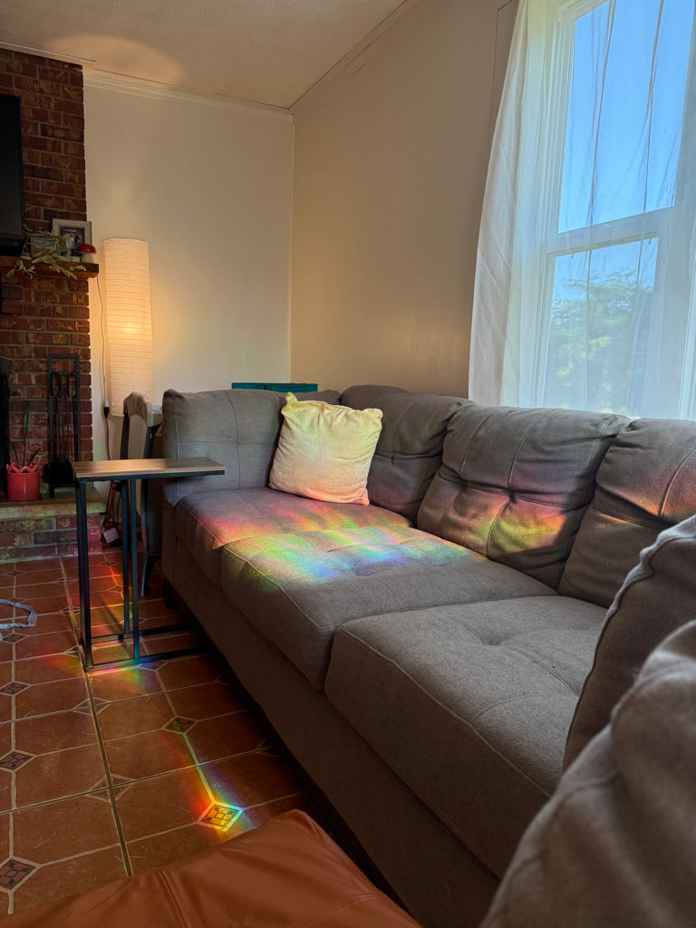 A gray fabric sectional sofa in a living room with a brick fireplace, tiled floor, and a window with sheer curtains. A colorful rainbow light projection is visible on the sofa and floor.
