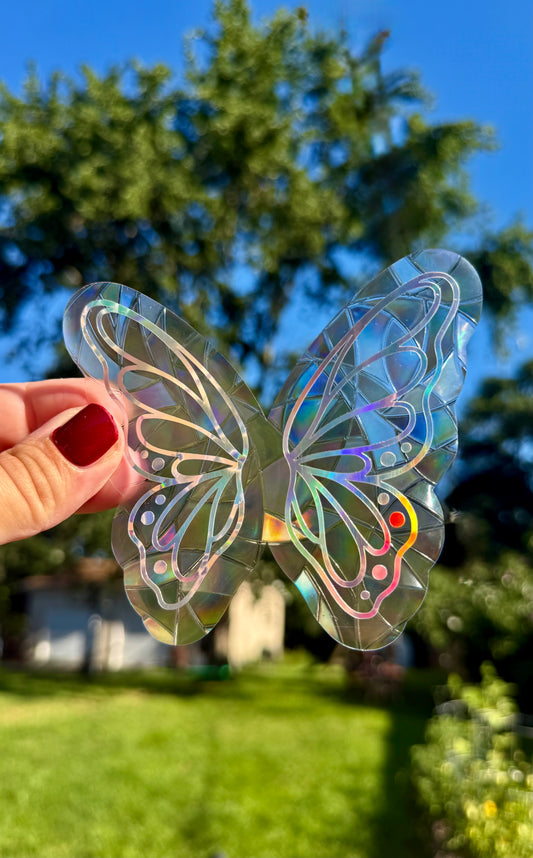 A hand with red nail polish holds a transparent butterfly-shaped decorative piece with holographic iridescent patterns against a blurred outdoor background of green grass, trees, and blue sky.