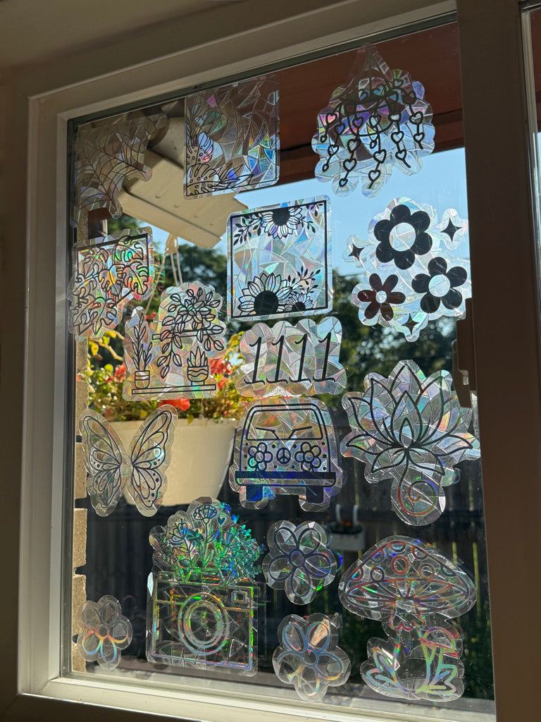 A collection of iridescent, holographic window decals featuring various designs including flowers, butterflies, a car, a camera, and mushrooms, arranged on a glass window.