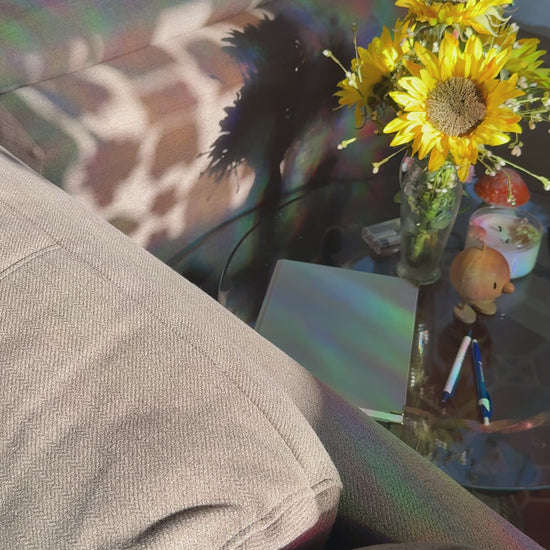 A cozy living room scene with a gray fabric sofa in the background, a glass coffee table in the foreground, and a vase of bright yellow sunflowers with rainbow light reflections on the table surface.