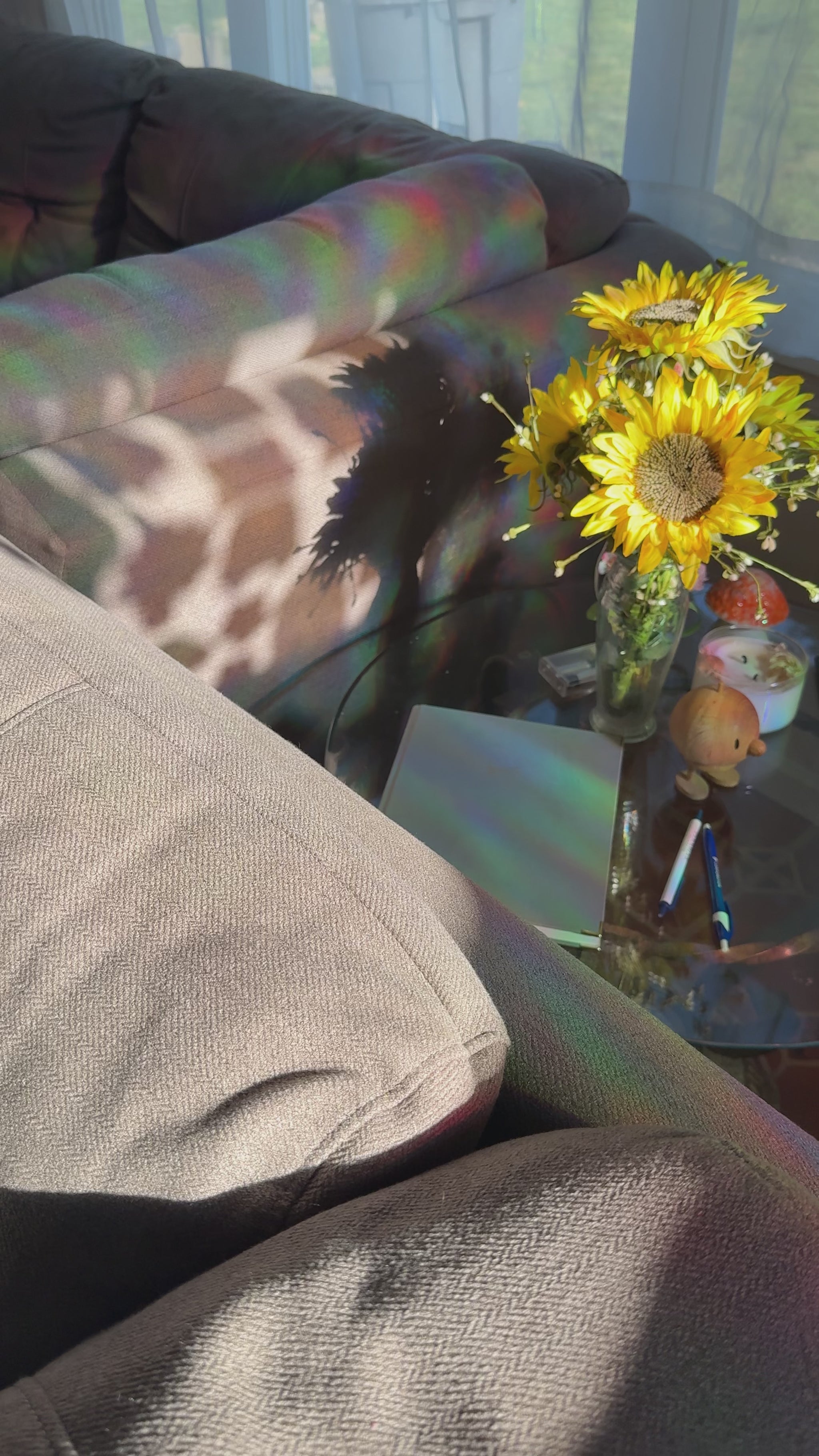 A cozy living room scene with a gray fabric sofa in the background, a glass coffee table in the foreground, and a vase of bright yellow sunflowers with rainbow light reflections on the table surface.