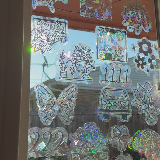 A collection of iridescent, holographic window decals featuring various designs including flowers, butterflies, a car, a camera, and mushrooms, arranged on a glass window.