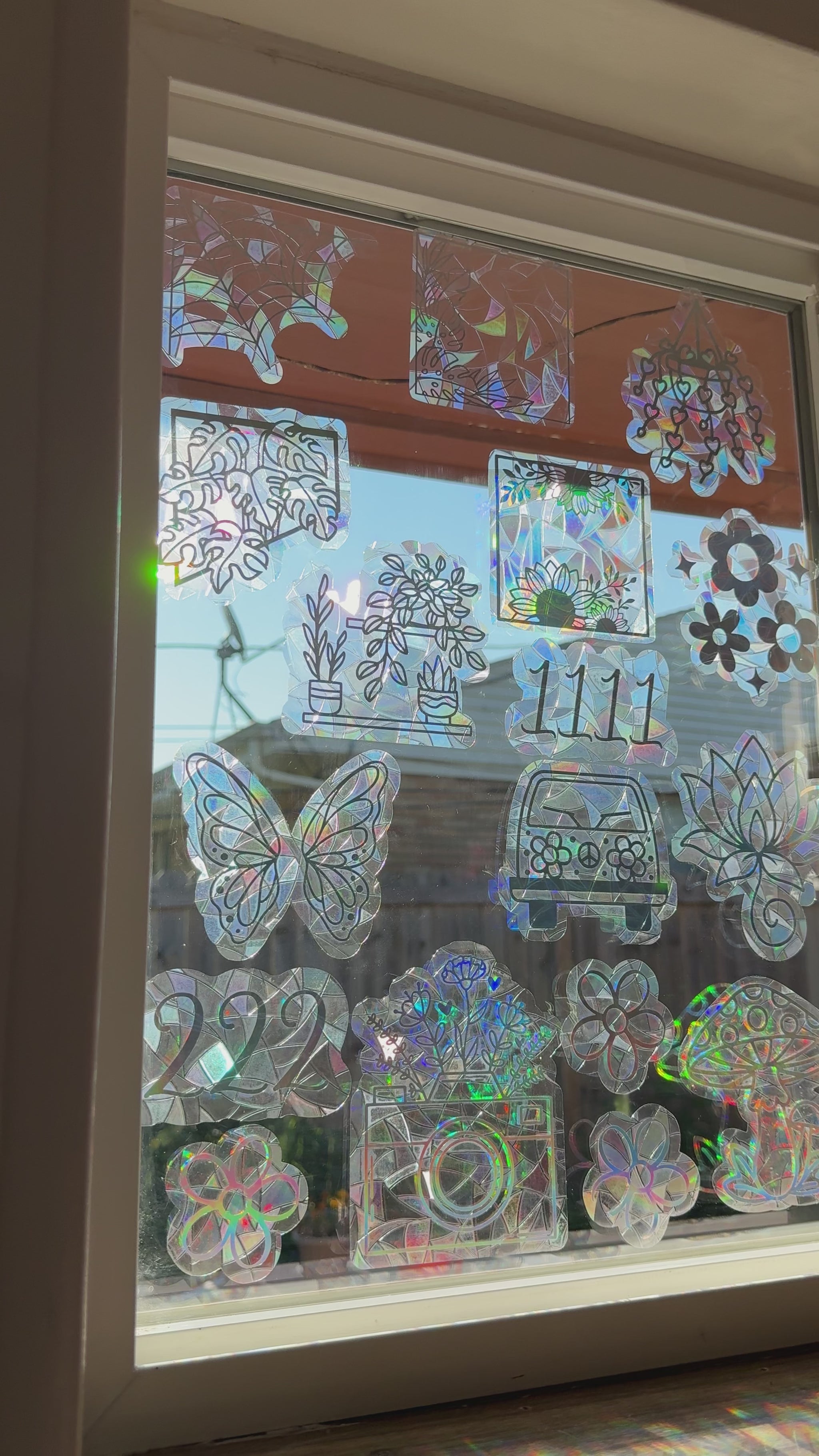 A collection of iridescent, holographic window decals featuring various designs including flowers, butterflies, a car, a camera, and mushrooms, arranged on a glass window.